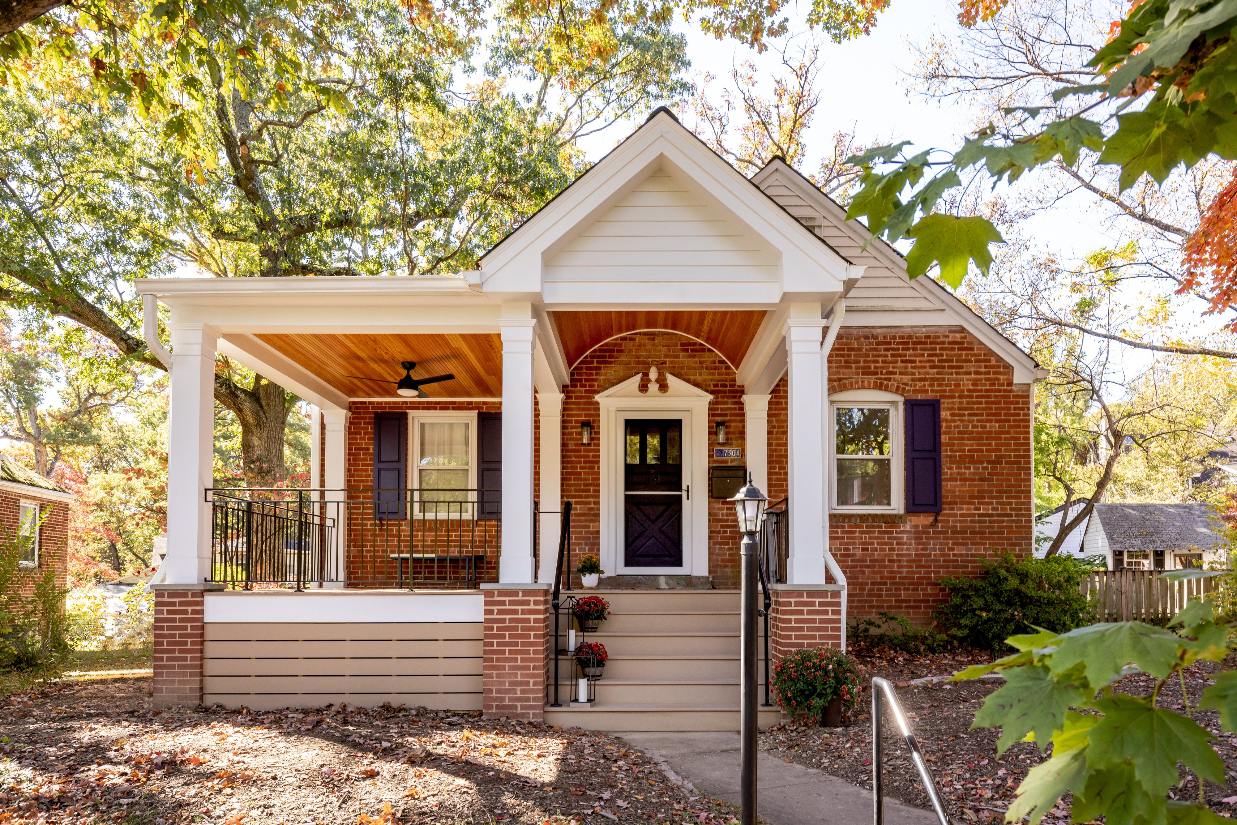 A cozy front porch with a wooden ceiling. It features a brick exterior, white pillars, and black railings. A cushioned chair with a pillow and a small plant sit near the door. Autumn foliage is visible in the background.