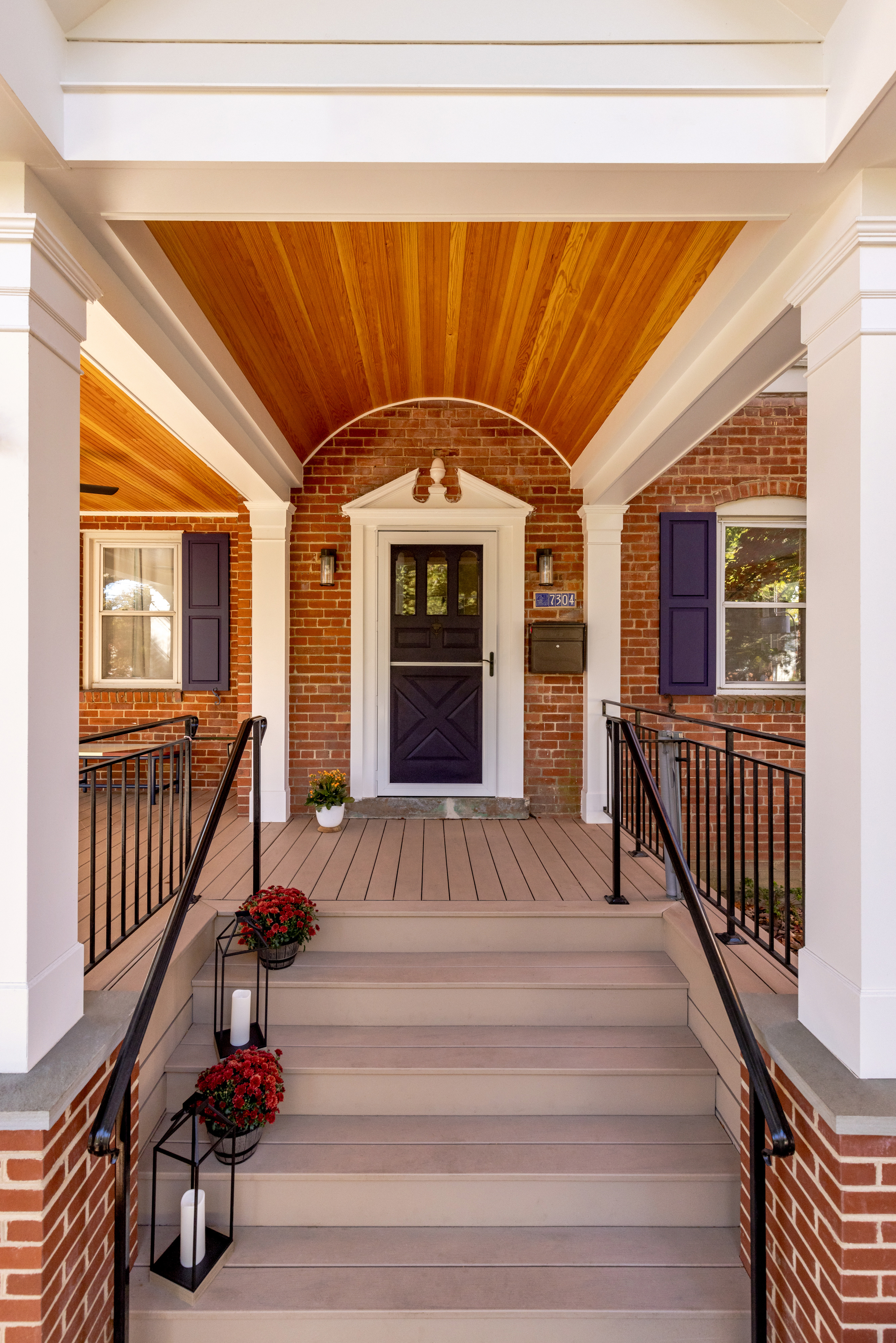 Trescott_New_FrontPorch17A house with a large front porch featuring a vaulted ceiling, white pillars and black metal railing, is surrounded by trees and plants, featuring windows and a front door, with siding that suggests it's in a historic style. The ground is covered with fall foliage, adding to the outdoor ambiance.