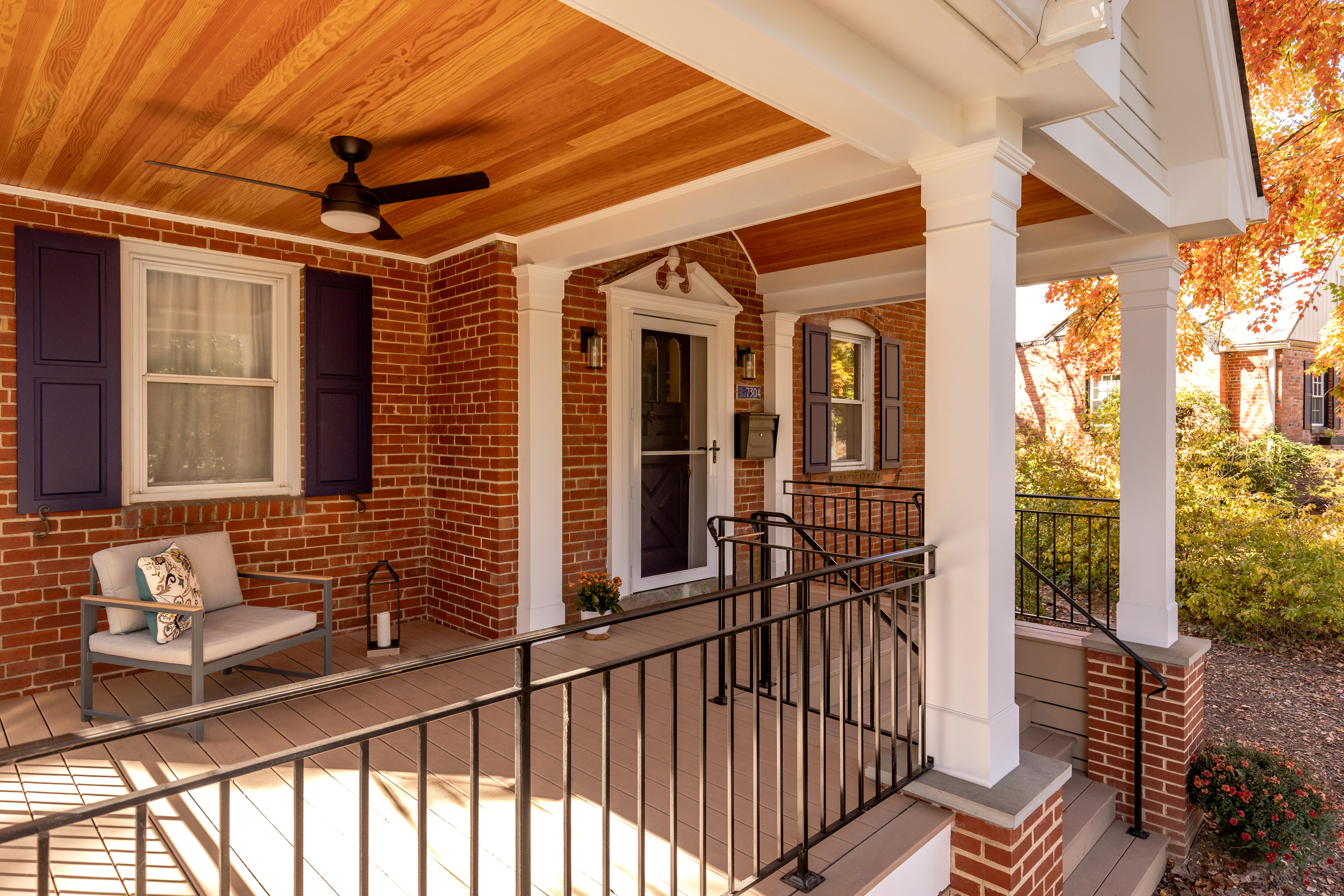 A house with a large front porch featuring a vaulted ceiling, white pillars and black metal railing, is surrounded by trees and plants, featuring windows and a front door, with siding that suggests it's in a historic style. The ground is covered with fall foliage, adding to the outdoor ambiance.