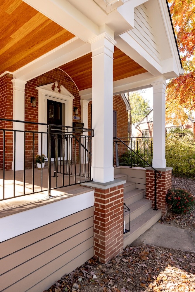 This Cabin John home's award-winning exterior features porch with a wooden ceiling and brick columns. Steps lead up to the entrance with a black door. Autumn leaves are scattered around, complementing the fall foliage of the surrounding trees.