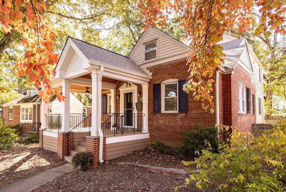 This Cabin John home's award-winning exterior features porch with a wooden ceiling and brick columns. Steps lead up to the entrance with a black door. Autumn leaves are scattered around, complementing the fall foliage of the surrounding trees. This Cabin John home's award-winning exterior features porch with a wooden ceiling and brick columns. Steps lead up to the entrance with a black door. Autumn leaves are scattered around, complementing the fall foliage of the surrounding trees.
