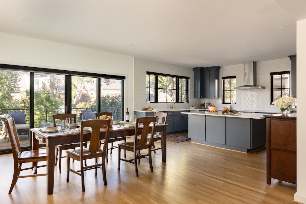 Modern kitchen and dining area in a Northwest DC home with wooden floors, a large dining table for six, gray cabinetry, white countertops, and large windows letting in natural light. Trees and houses are visible outside.