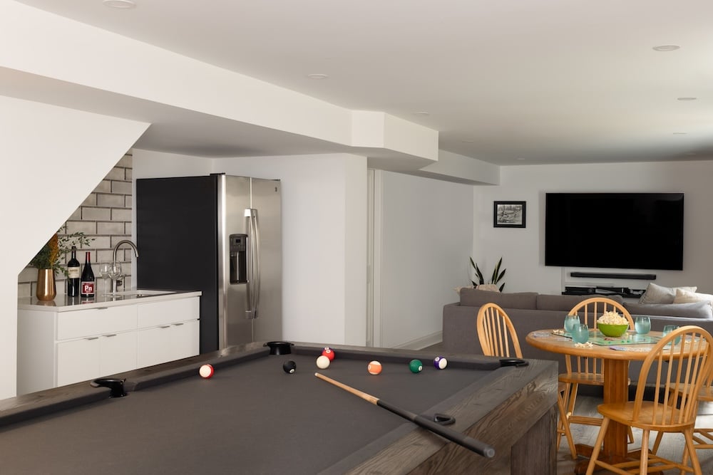 A modern basement recreation room by a Northwest DC award-winning remodeler, featuring a black pool table, round dining table with wooden chairs, gray sofa, wall-mounted TV, mini fridge, and a small kitchenette—a stunning custom-remodeling project.